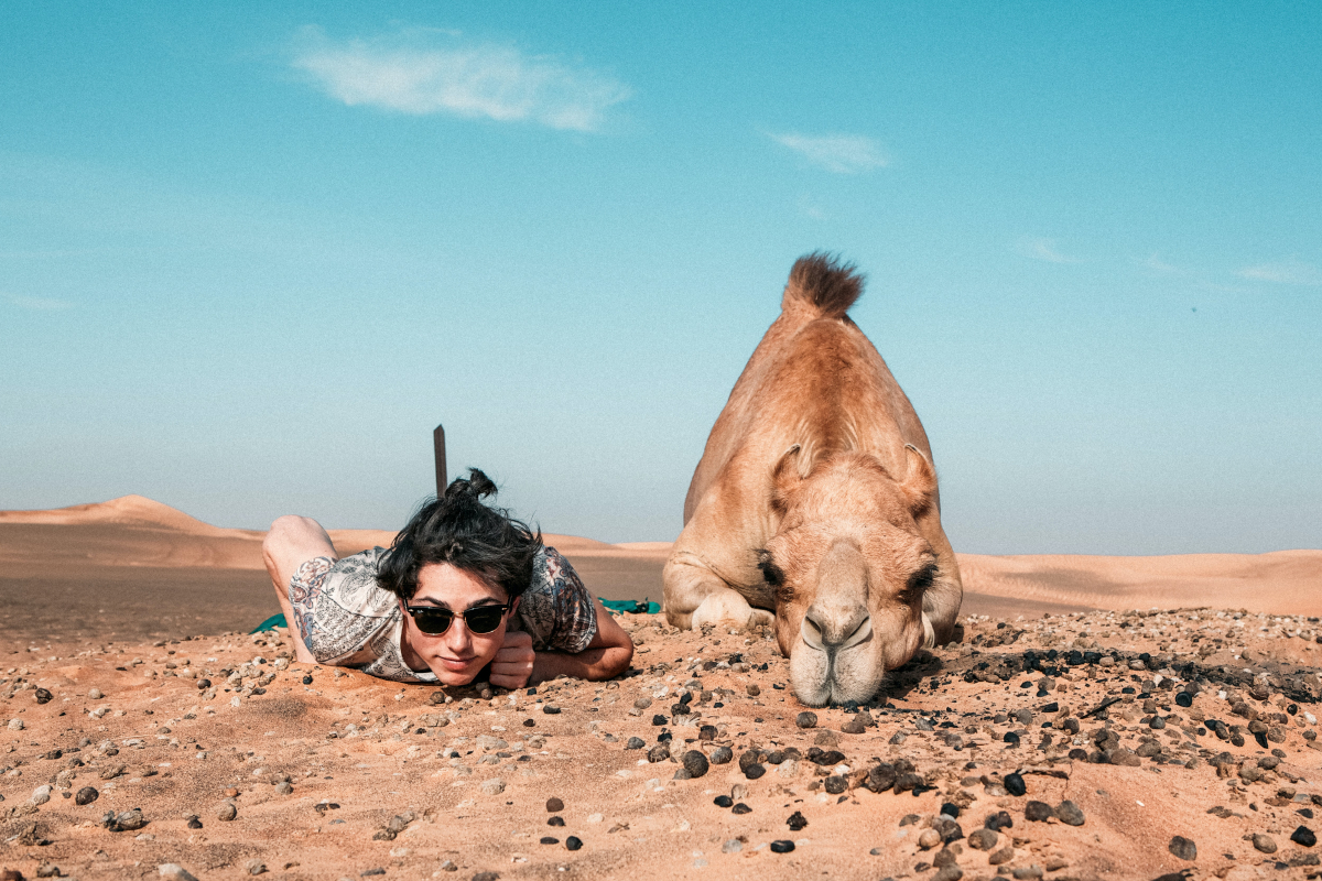 Mann und Kamel liegen in der Wüste auf dem Boden, gucken in die Kamera